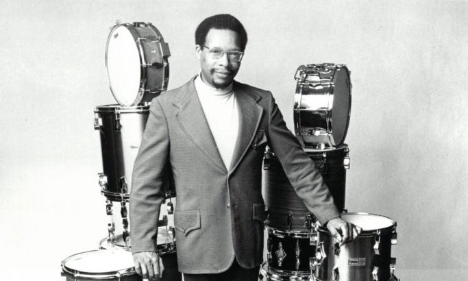 Black and white portrait of Alan Dawson standing in front of drums.
